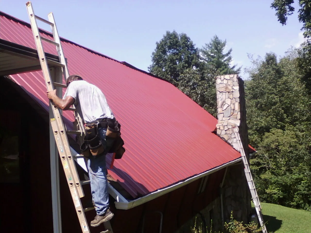 Roofer on a ladder beside a red metal roof and stone chimney in Paoli